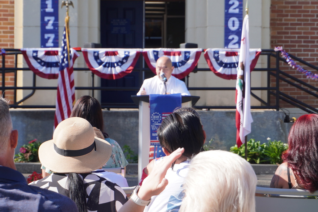 A person speaks from a podium with flags in front. Several people sit behind them, facing the speaker.