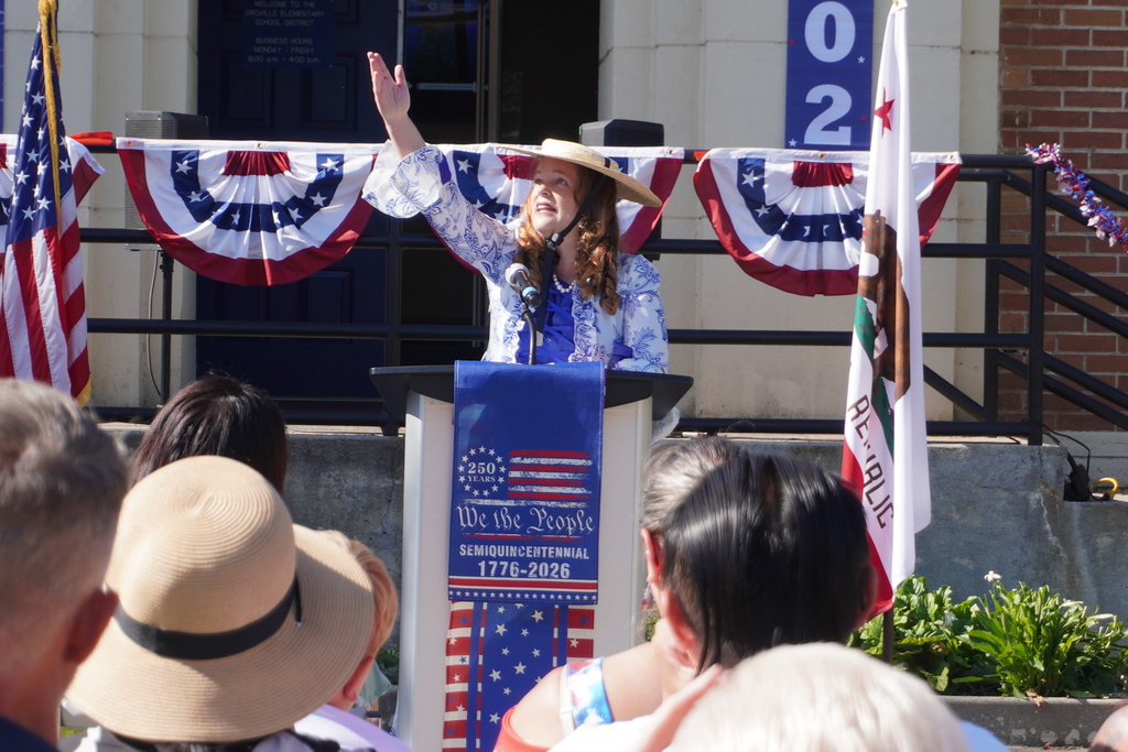 A person stands at a podium, speaking into a microphone, with an audience seated before them.