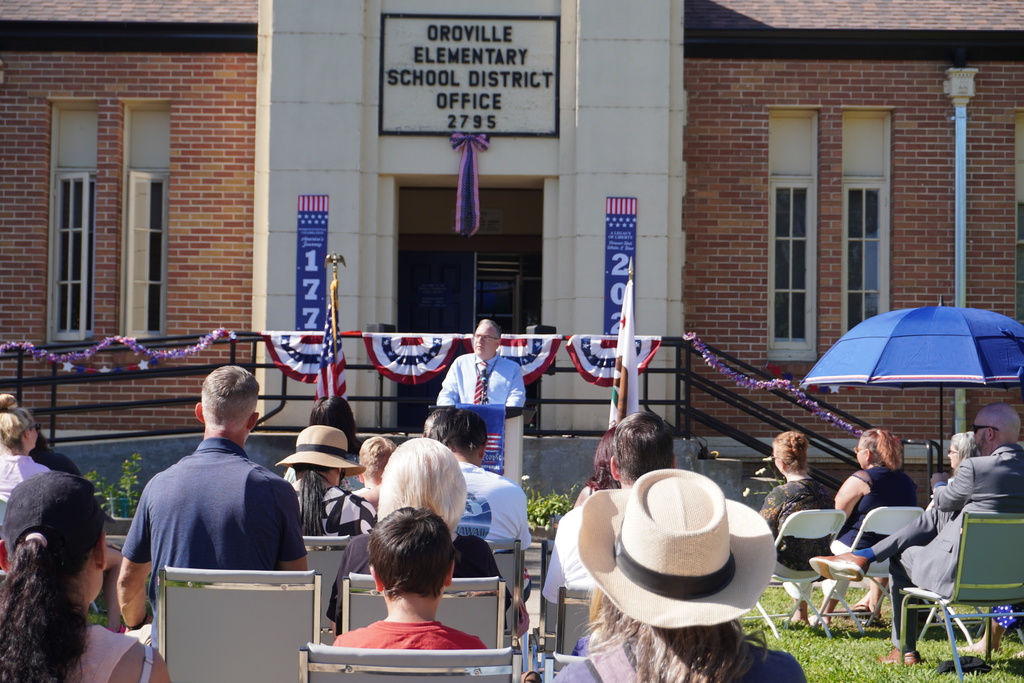 People seated in chairs around a stage. A man speaks at a podium. Behind him is the Groville Elementary School District Office.