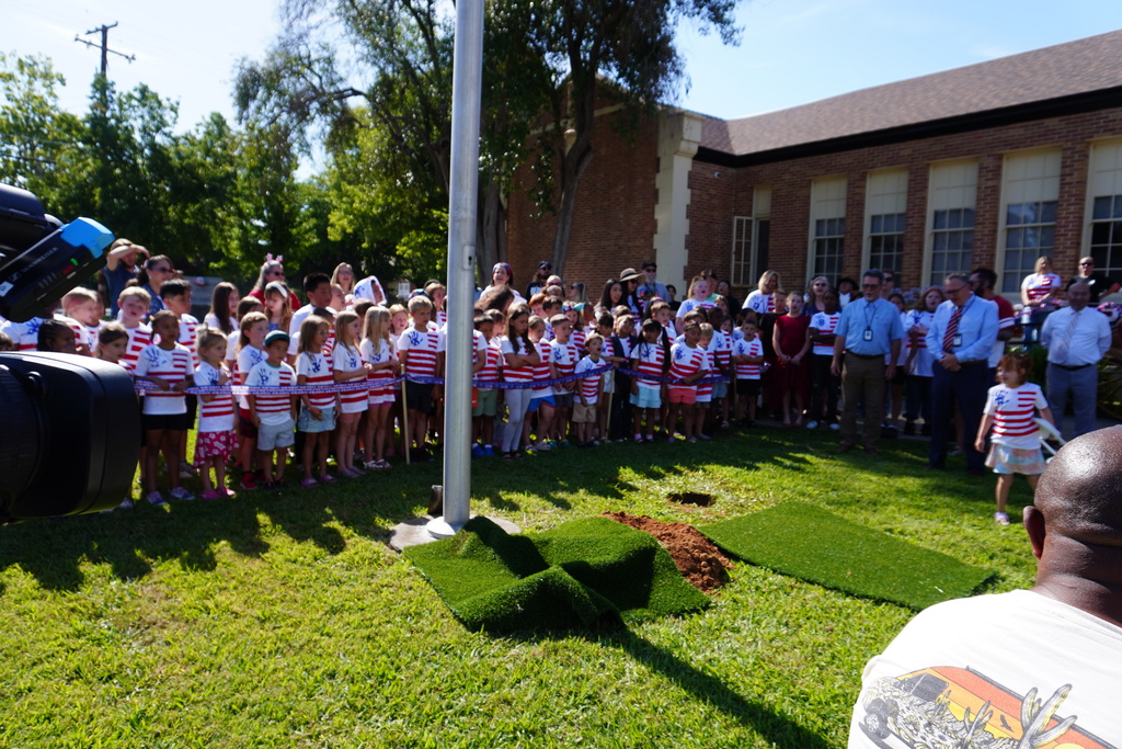 Large crowd standing on grass next to a pole. Many children in white shirts and shorts. Adults in suits.