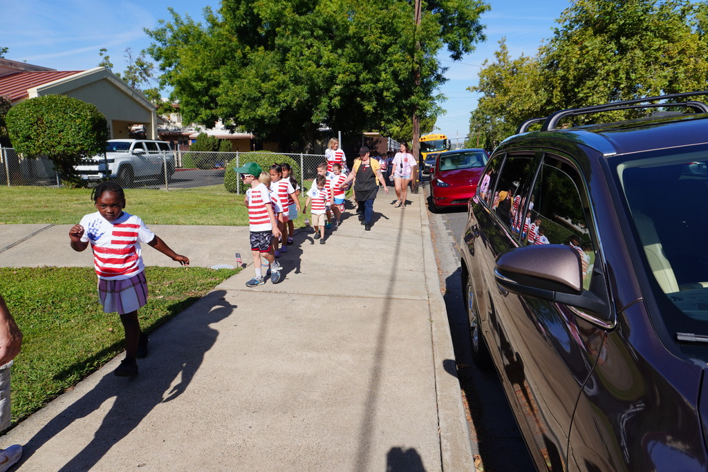 Sidewalk with children wearing striped shirts, a parked car, and houses in the background.