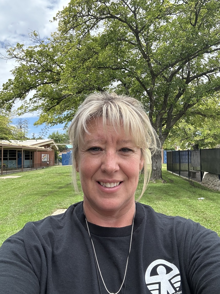 A woman with blonde hair stands outdoors, smiling. A white logo is on her black shirt. Behind her is a tree, fence, and building.