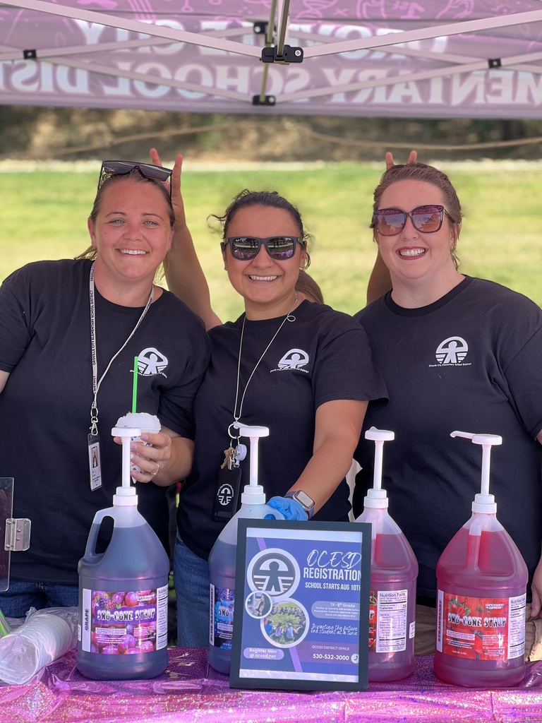 Four women in black shirts and sunglasses stand behind a table with bottles of purple liquid and a plaque.