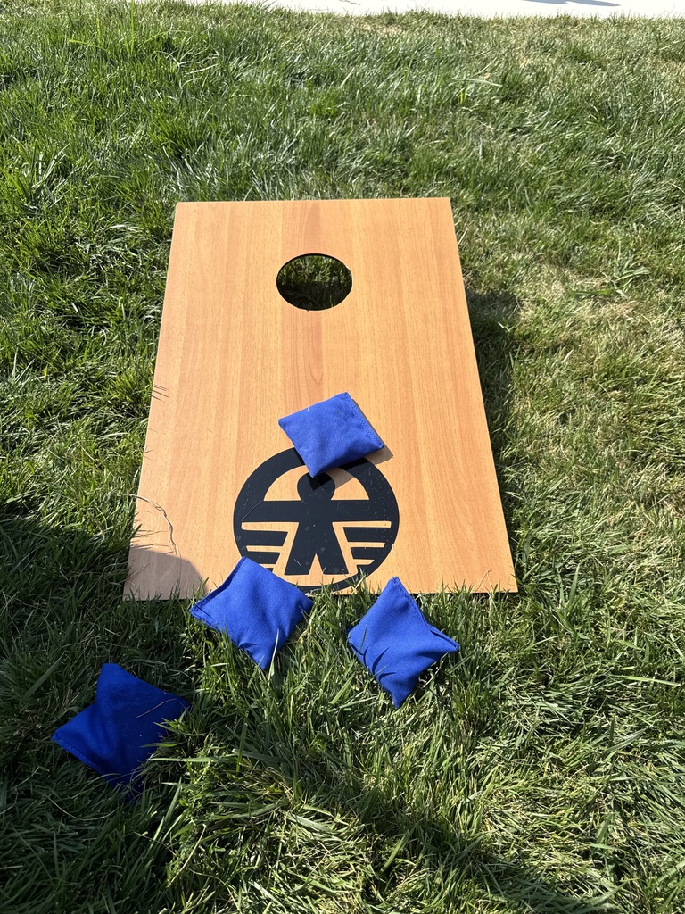 A wooden cornhole board with four blue bean bags nearby on a green lawn. The board has a hole in the center.