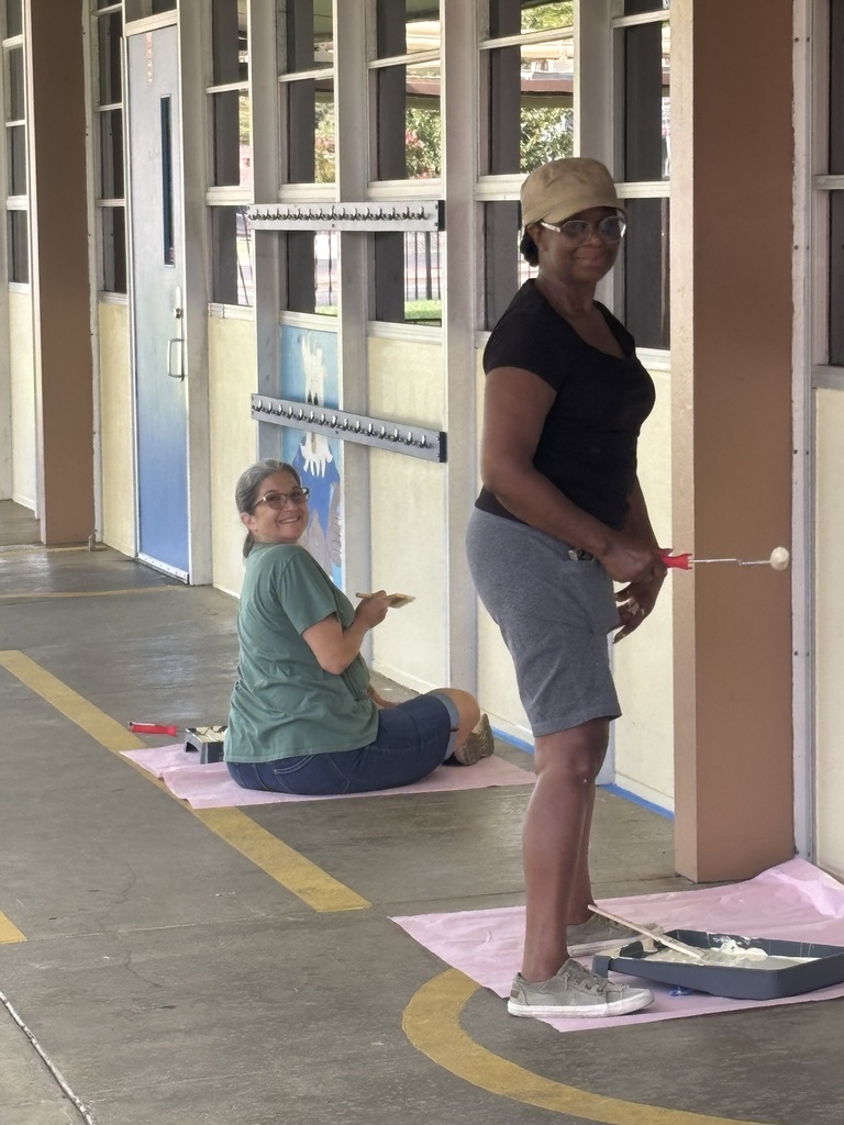 Two women wearing glasses are painting a wall with rollers. One sits on a mat and the other stands.