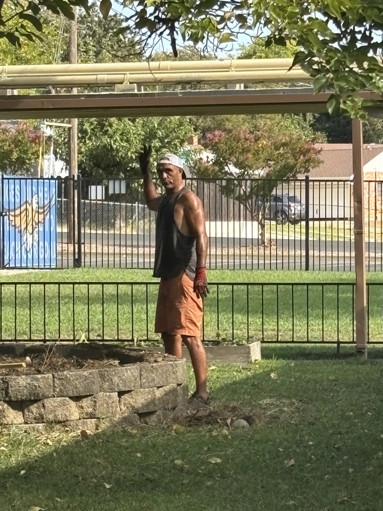 Person in orange shorts and a hat stands in a park near a fence, waving.