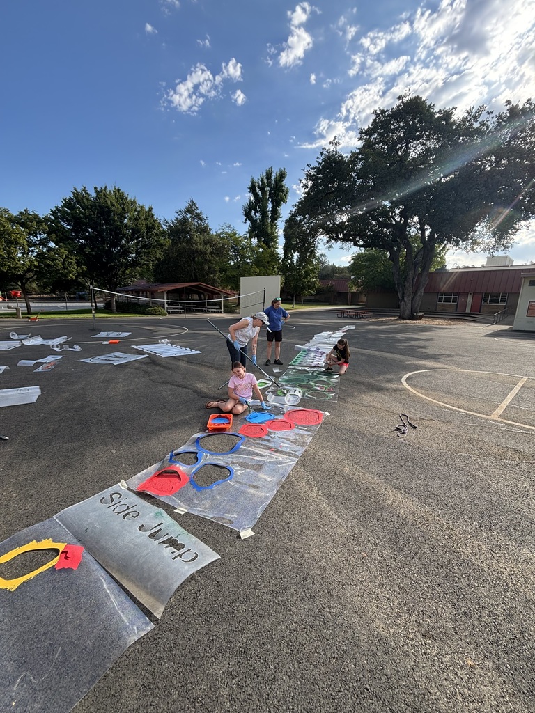 A group of people are painting on a large canvas laid out on a paved surface, with a blue sky and trees in the background.