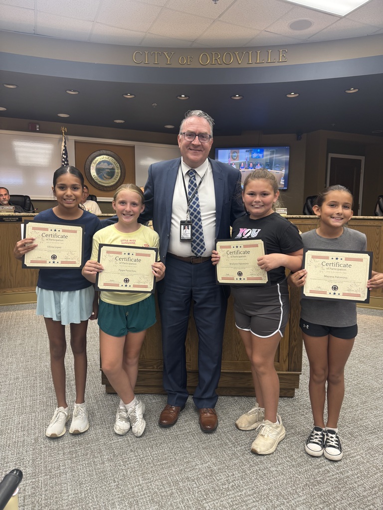 Five young girls hold certificates in a city council room. An adult man in a suit and tie stands between two of them.