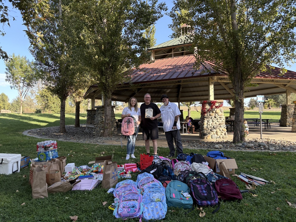 Three people stand in a park with bags and boxes around them. A shelter with a red roof is behind them.
