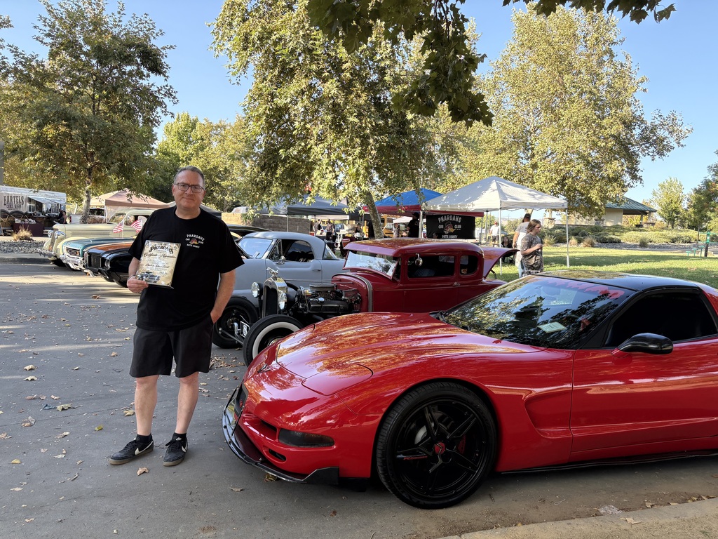 Person holds award near a red sports car at an outdoor auto show with tents and trees.