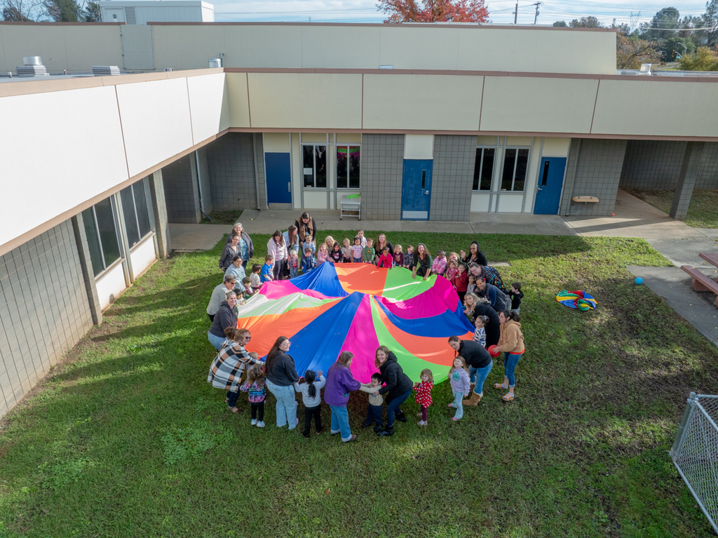 Group gathered around a colorful parachute, in a yard. Behind them is a building with blue doors.