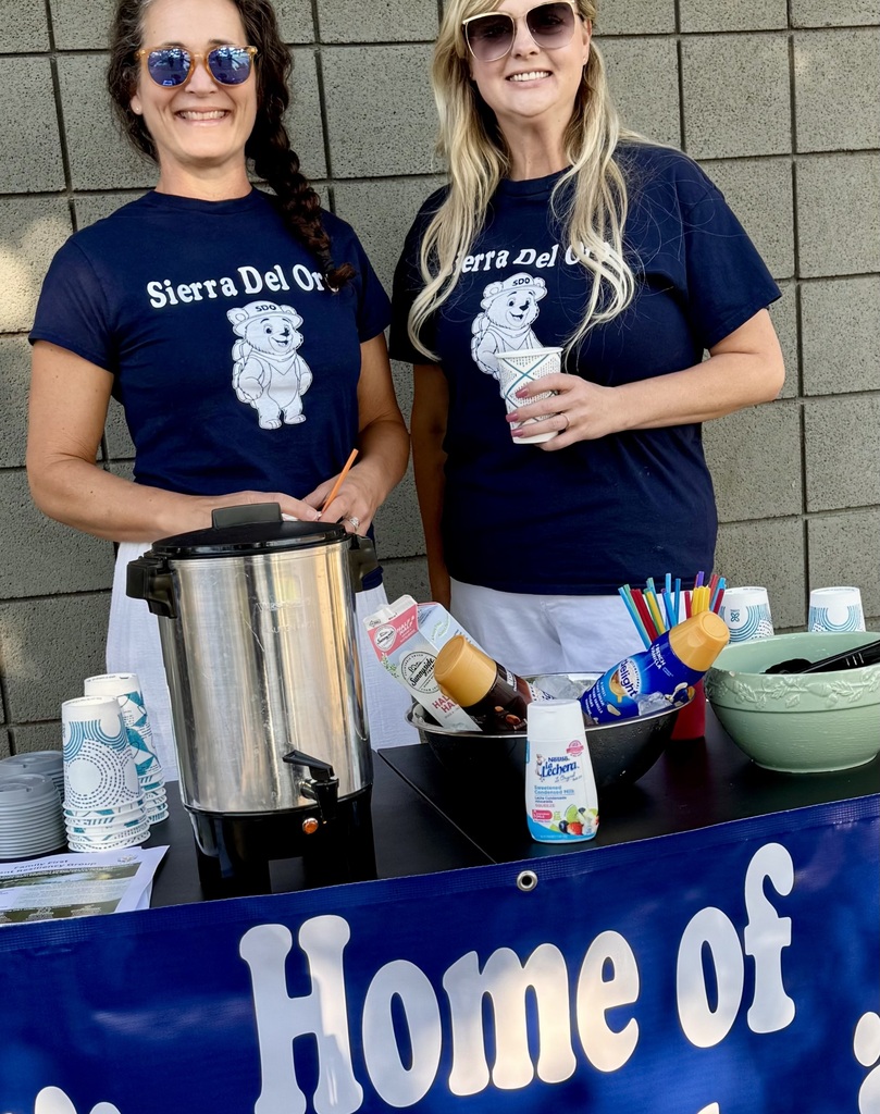 Two women stand at a table, smiling and wearing blue shirts. A banner reads "Home of". Coffee and drinks are on the table.