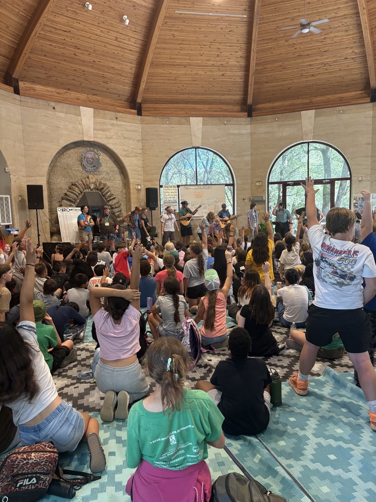 A group of people gathered in a round room with wooden ceiling, some seated and others standing, with musical instruments present.
