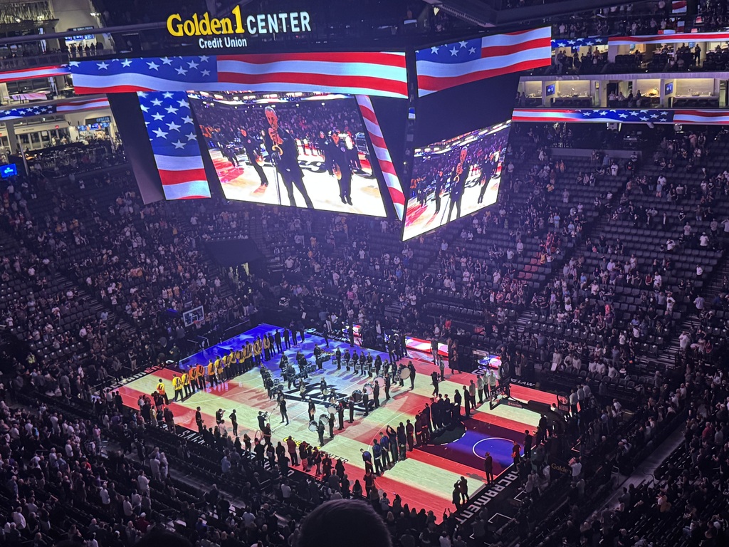 Large indoor stadium with packed stands, large screens, and a basketball court with players in a circle.