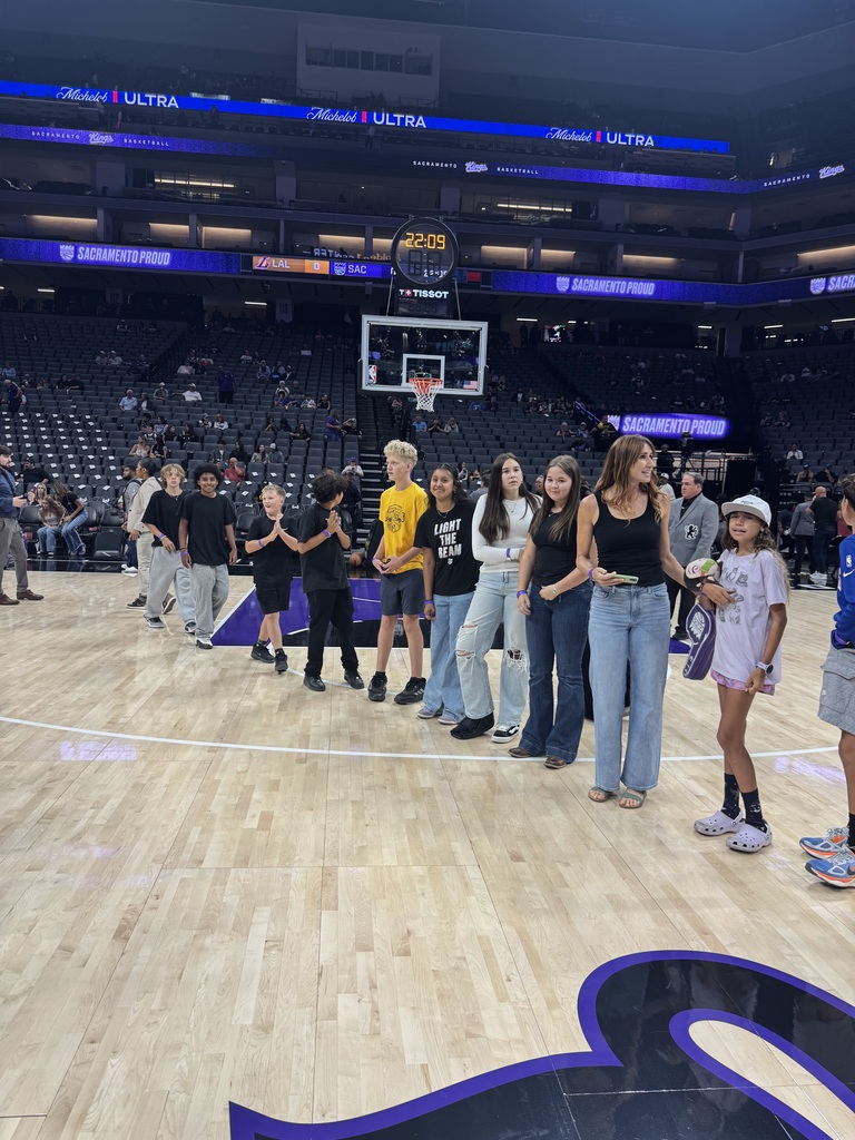 A group of people stand on a basketball court, near a hoop. The floor has blue markings. Advertisements and empty seats are in the background.