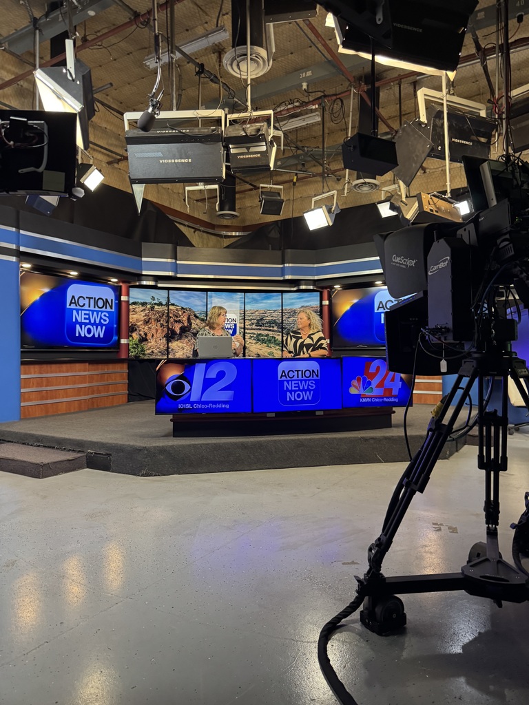 Studio floor with a blue LED news screen, cameras, and lights. Blue walls and white flooring. A camera on a tripod is positioned in the foreground.