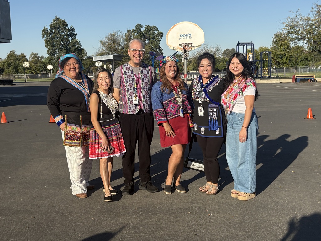 Seven people wearing traditional clothing stand in a parking lot with a basketball court in the background.
