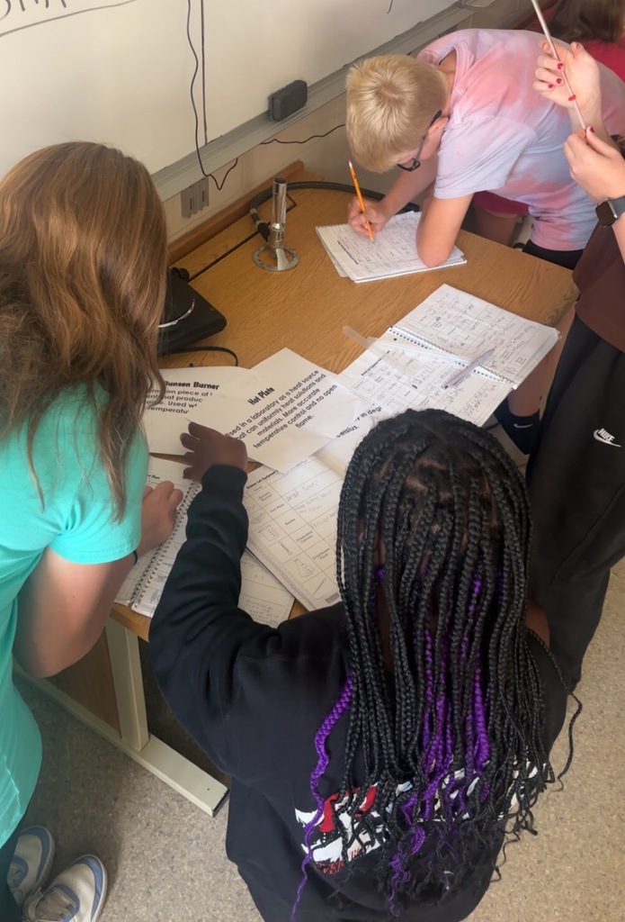 Image Description for accessibility: Students looking a science equipment and writing in notebooks