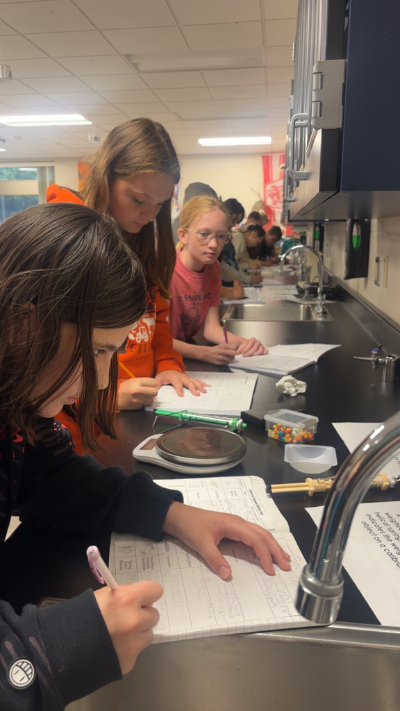 Image Description for accessibility: Students looking a science equipment and writing in notebooks