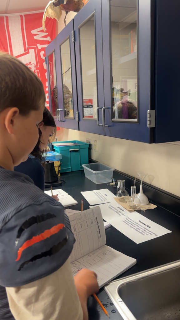 Image Description for accessibility: Students looking a science equipment and writing in notebooks