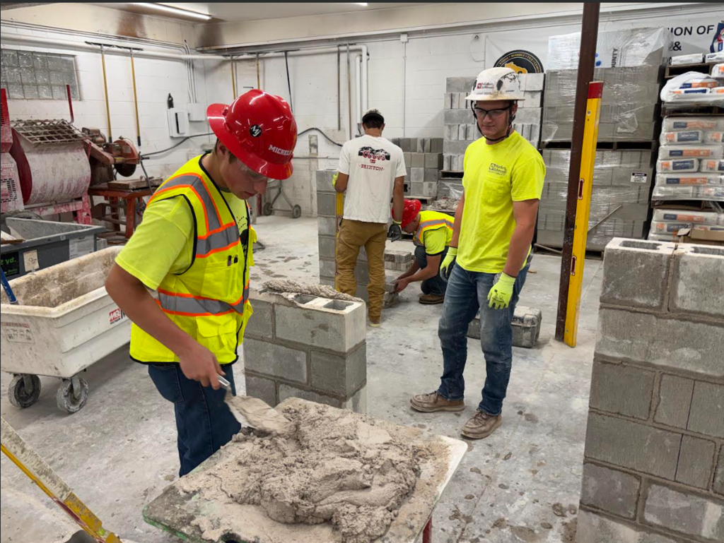 Image Description for accessibility: Student wearing reflective vest and hard hat practicing brick laying techniques