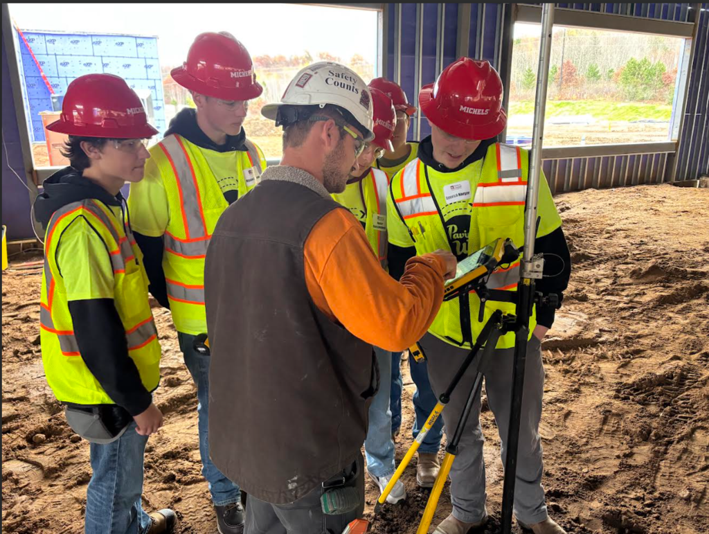 Image Description for accessibility: Students in reflective vests and hard hats watching a demonstration