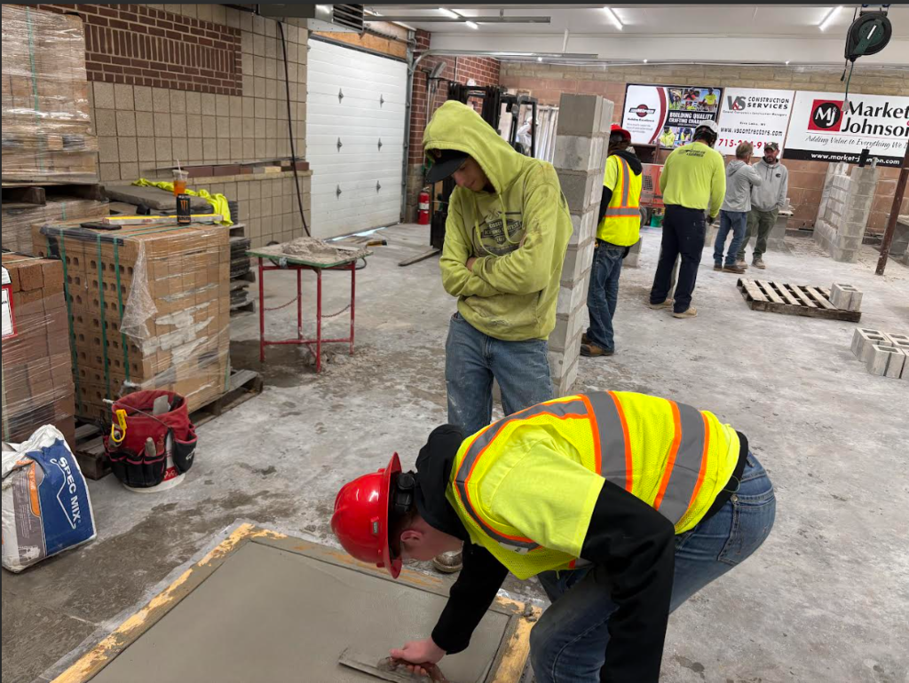 Image Description for accessibility: Students in reflective vests and hard hats smoothing wet concrete