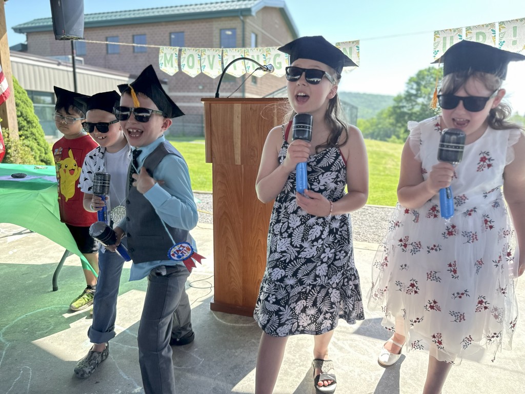5 elementary aged students in graduation caps and sunglasses with microphones singing