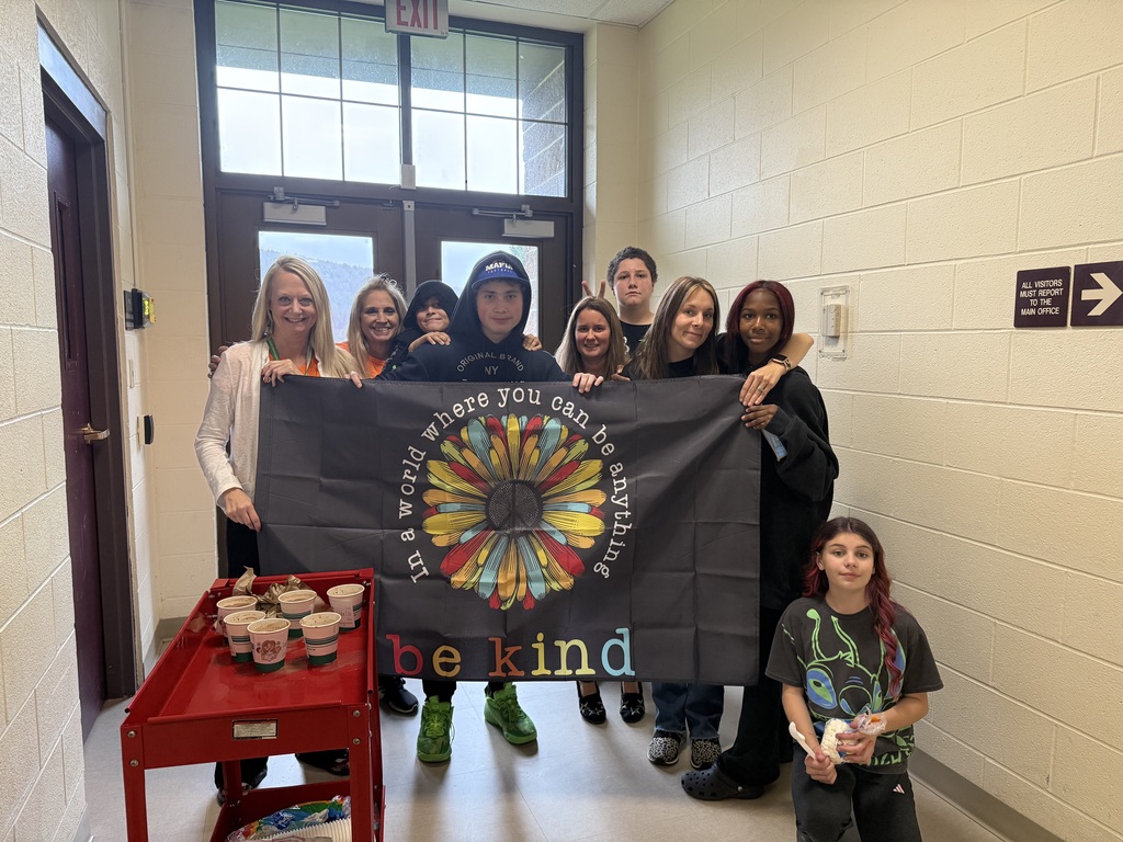 Students and staff with a cart of hot cocoa and a sign saying "in a world where you can be anything be kind"