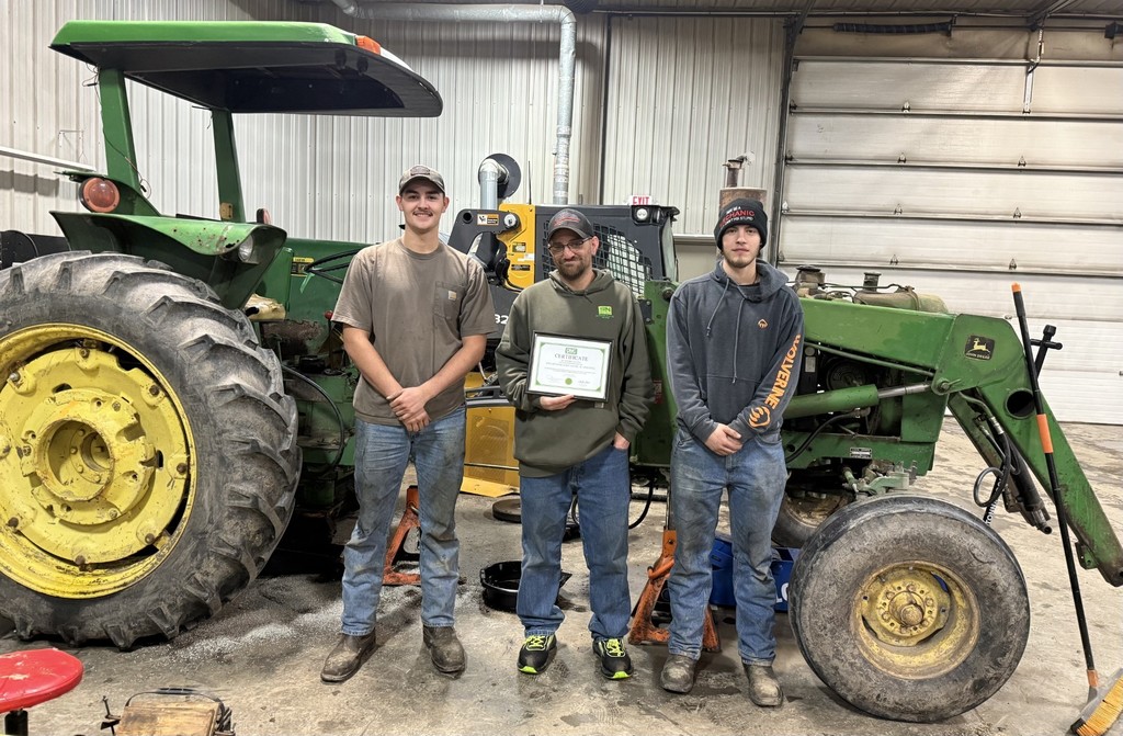 Two students and SN John Deere employee standing in front of John Deere tractor with a certificate