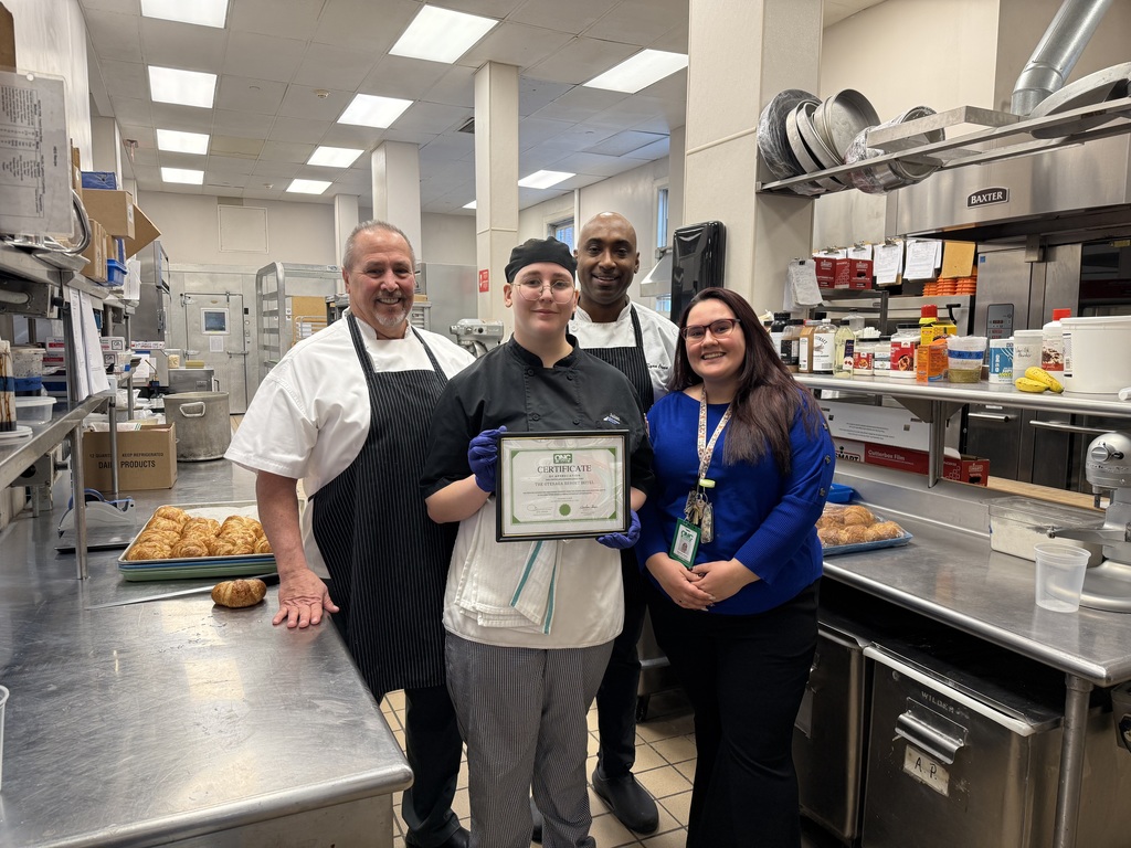 Student in a restaurant kitchen holding a certificate with the culinary arts instructor and two chefs