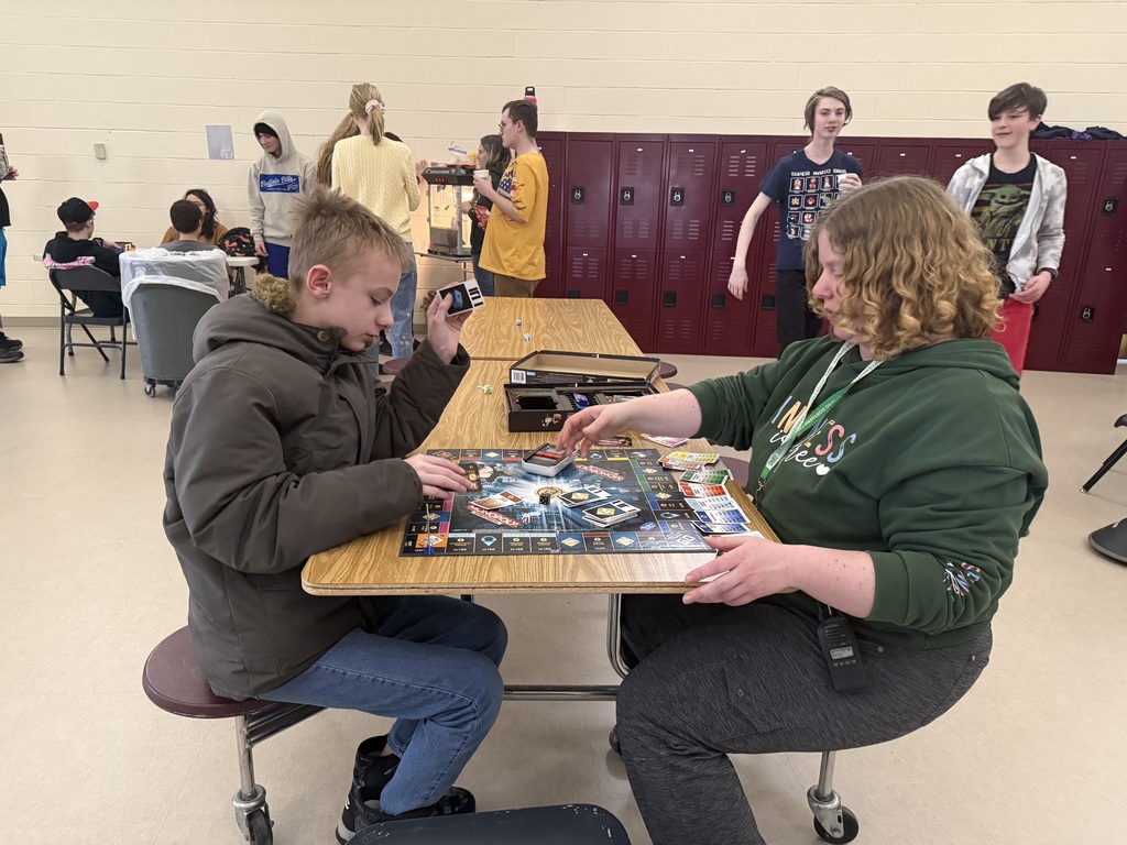 Two students sitting at a table playing a board game with other students in the background
