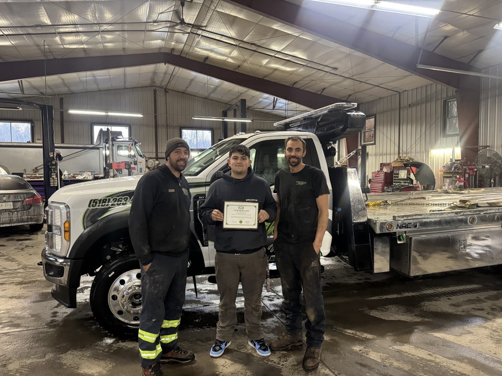 Student and two workers of Pete’s Auto Repair and Towing standing in front of a tow truck inside a garage.