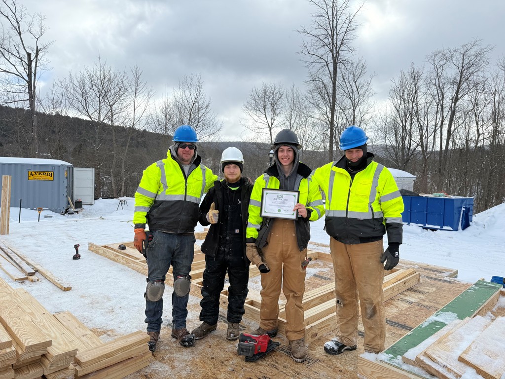 Two students, one holding a certificate, with two workers from Eastman Associates. All outside with heavy jackets and hard hats.