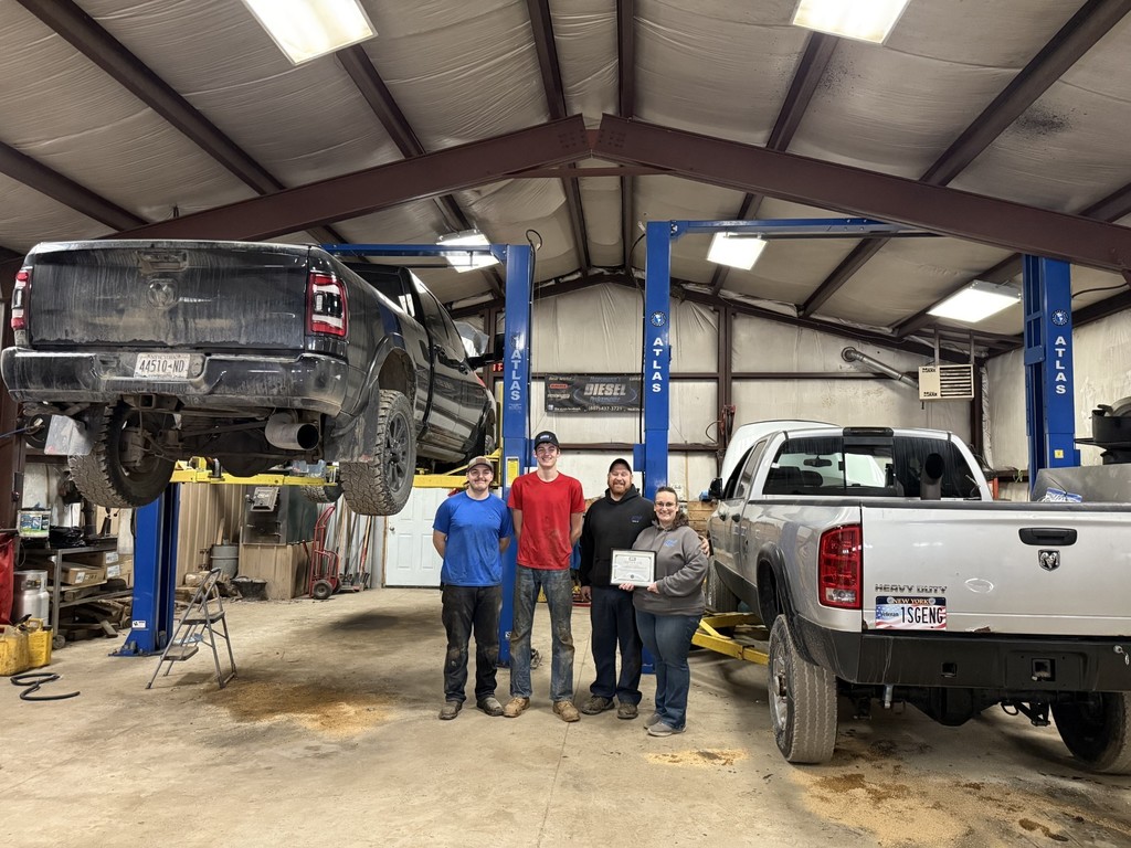 Two students and two employees of Hausmann's Diesel Performance LLC standing in an auto shop with two trucks, one up on a lift, holding a completion certificate.