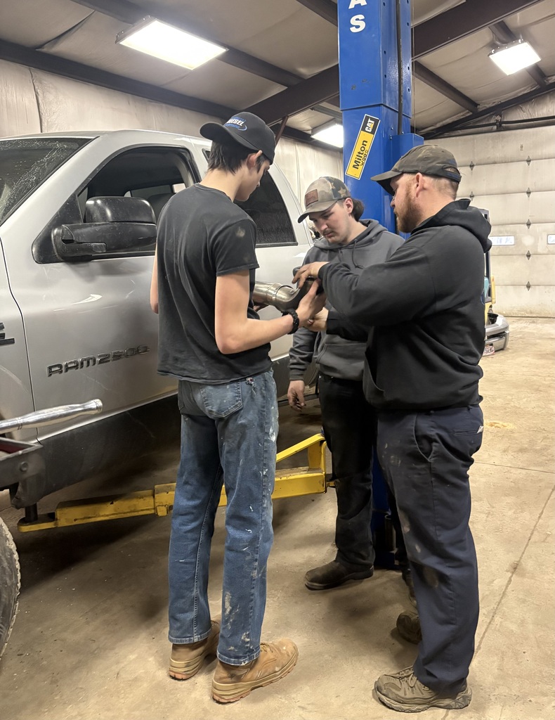Two students and employee standing by a truck. One student is holding a vehicle part and the employee is explaining something.
