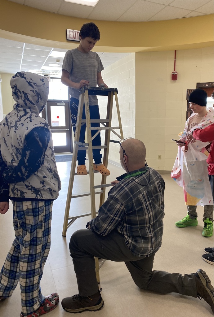 Three students, one on a ladder, with a teacher kneeling in front of the ladder holding it.