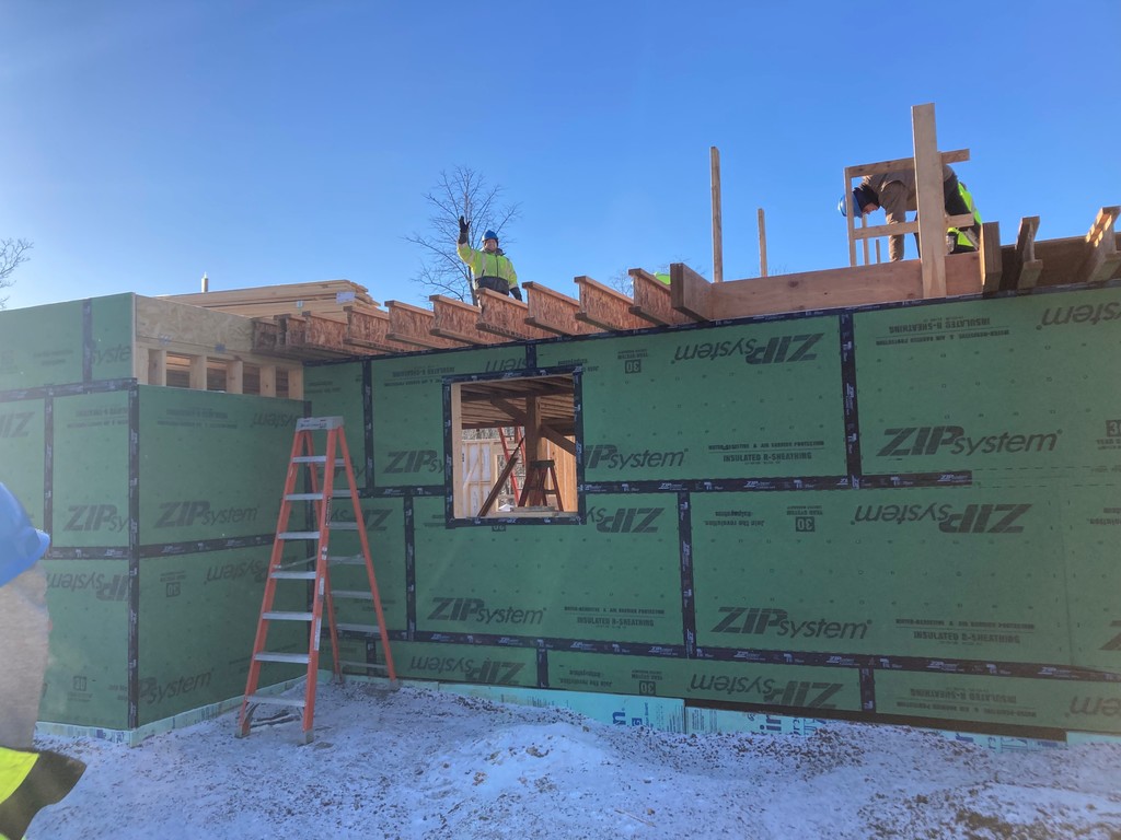 Student on top of a building under construction waving. Another worker is off to the side. A ladder is in front of the building being built.
