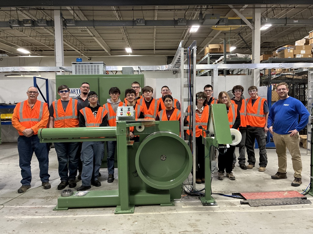 13 students and the instructor in orange safety vests along with two employyes. They are standing behind a large green piece of machinery.