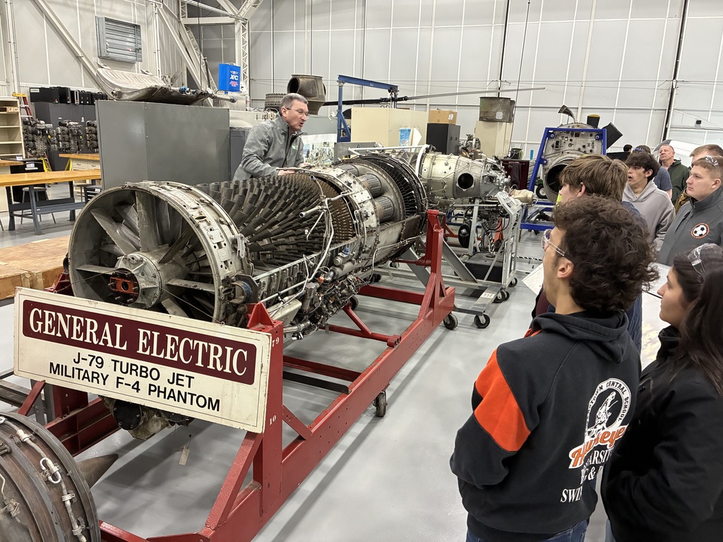 Students listening to an employee speak. They are standing near an engine that is labeled General Electric J-79 Turbo Jet Military F-4 Phantom