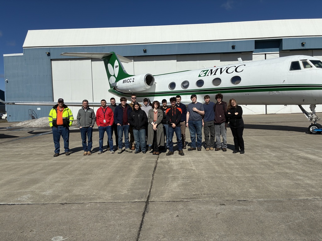 Thirteen students, class instructor, and two employees standing on a runway in front of a white and green airplane that says MVCC.