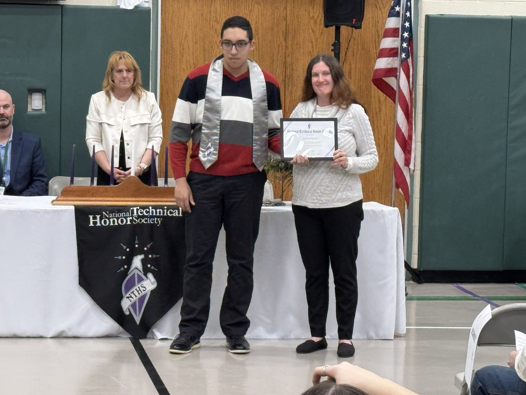 An instructor and a newly inducted student posing in front of a table with a black National Technical Honor Society banner. Two staff members are in the background.