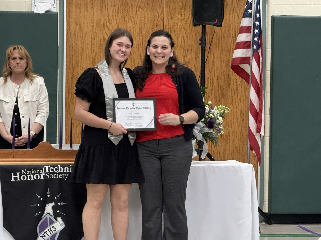 An instructor and a newly inducted student posing and smiling in front of a table with flowers and the American flag