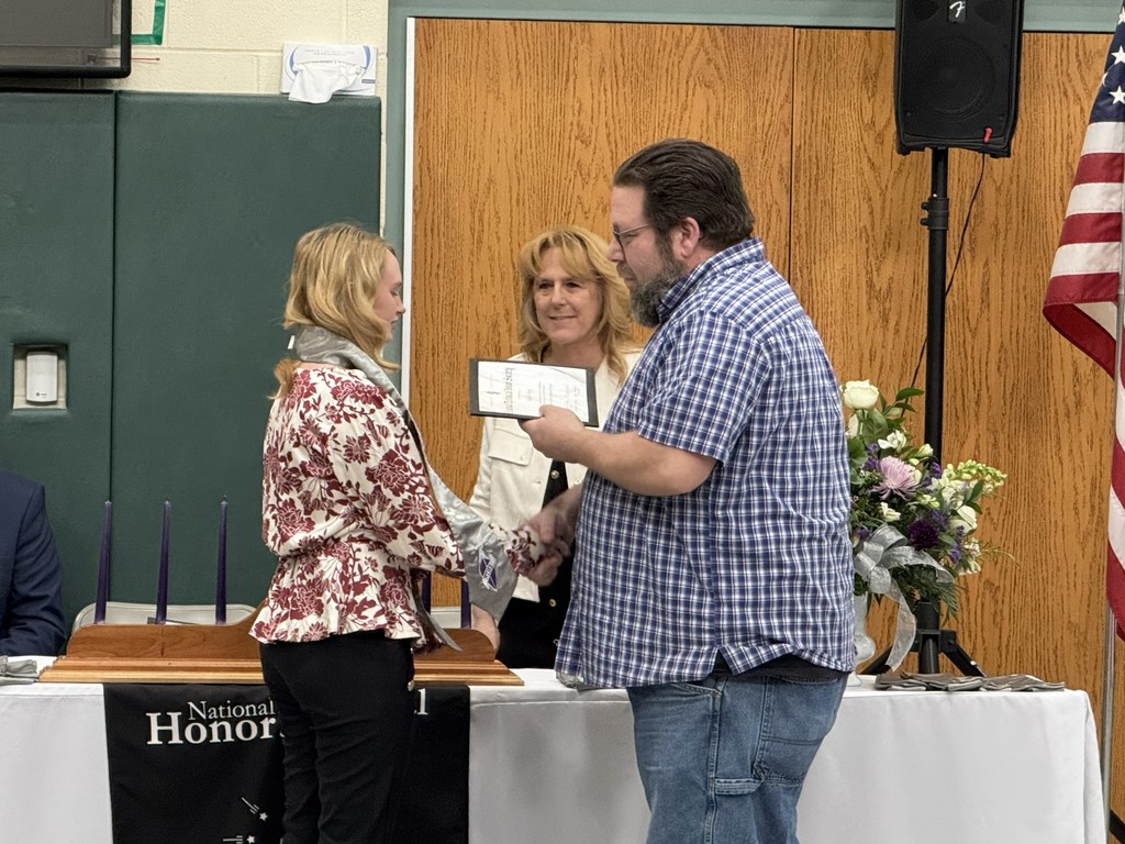 An instructor shaking the hand of a newly inducted student and holding a certificate. There is a staff member in the background.