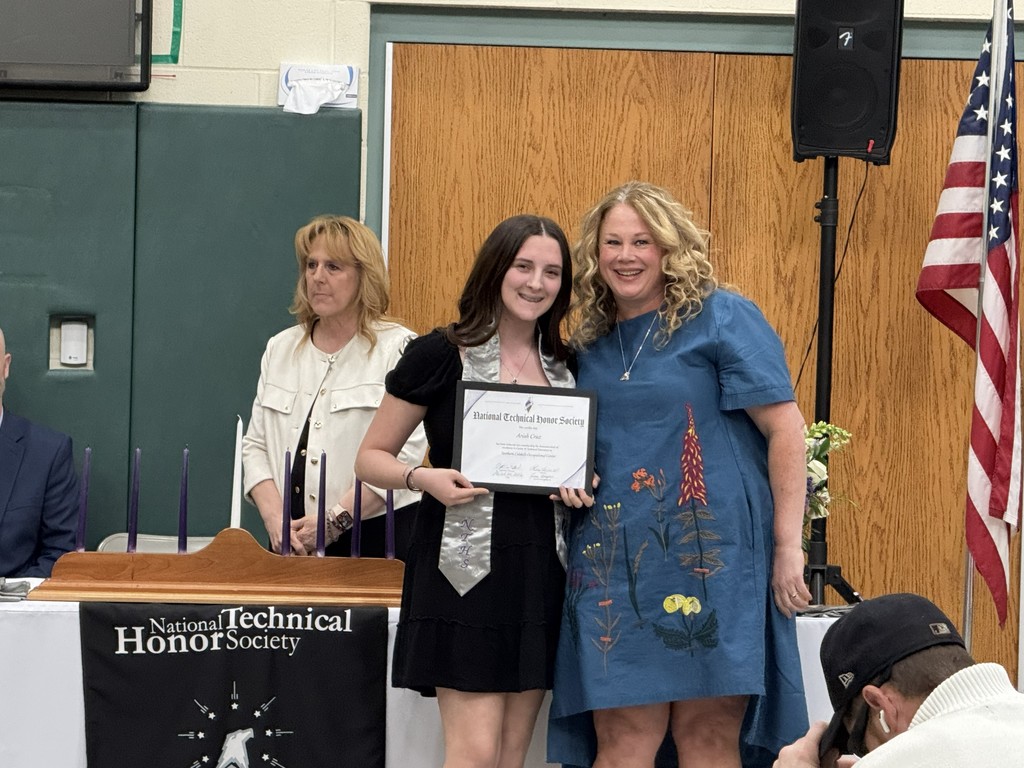 A newly inducted student holding a cerificate is posing with their instructor. There is a NTHS black banner to the one side and the American flag on the other side. There is a staff member in the background.