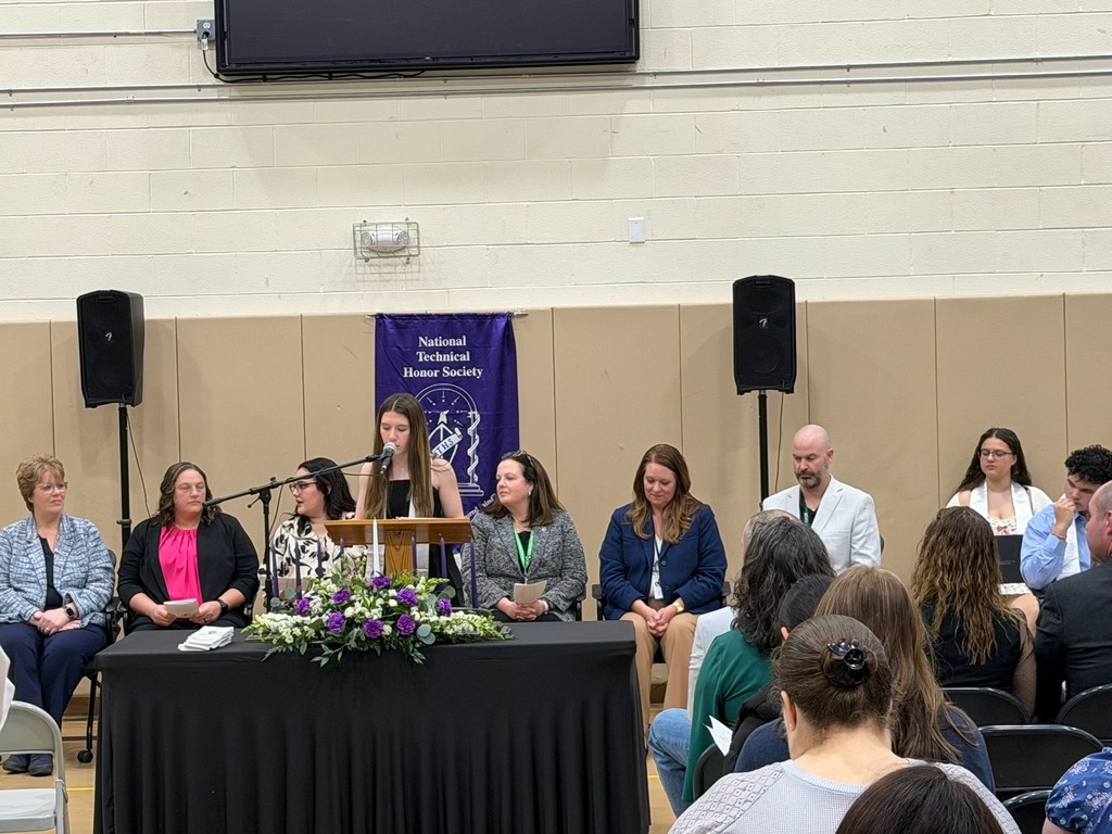 Student is speaking at a podium with a microphone. There is a black tablecloth and flowers on the table. There are staff members sitting behind the student.