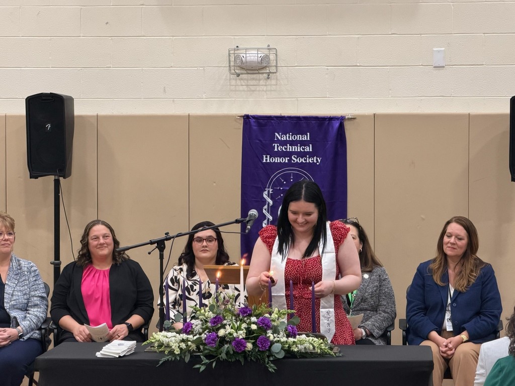 A student lighting a candle representing the pillars of NTHS. There is a purple NTHS banner hanging behind the student and staff members sitting.