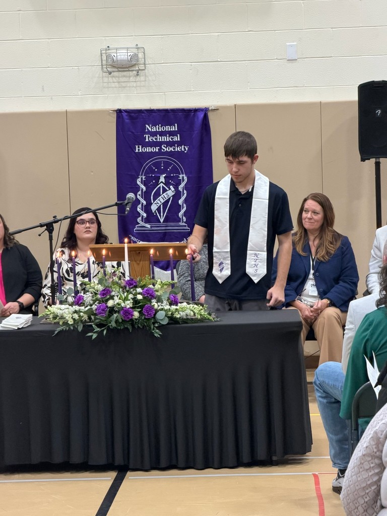 Student lighting a candle representing the pillars of NTHS. There is a purple NTHS banner hanging on the wall behind the student and staff members sitting behind the student.