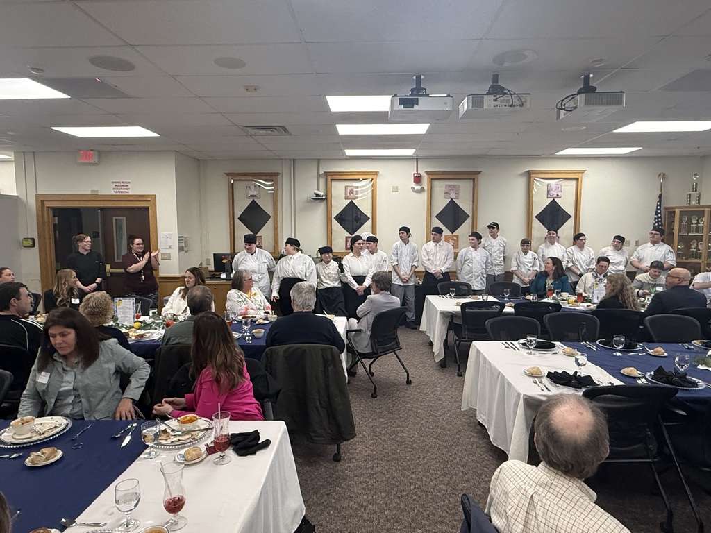 Attendees sitting at tables in the dining room acknowledging the culinary students who are standing.