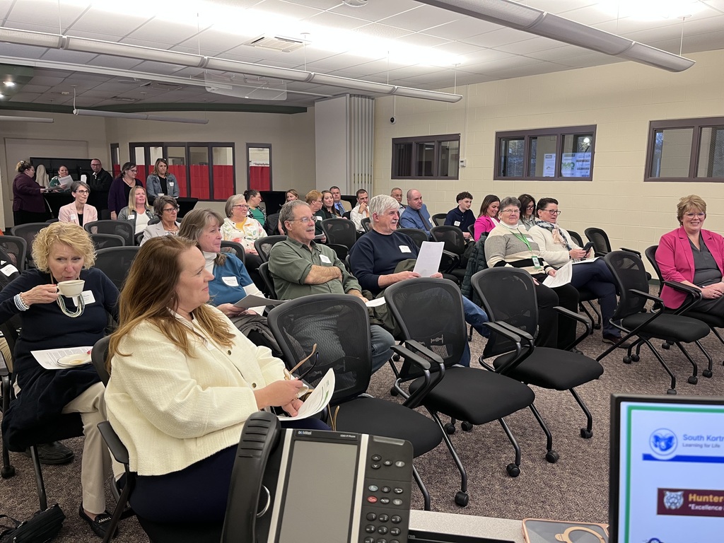 A  picture of the audience at the meeting sitting in chairs and standing in the back of the room